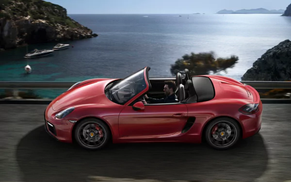 Red Porsche Boxster GTS convertible cruising a coastal road with rocky cliffs and calm sea — HD PC desktop wallpaper background.