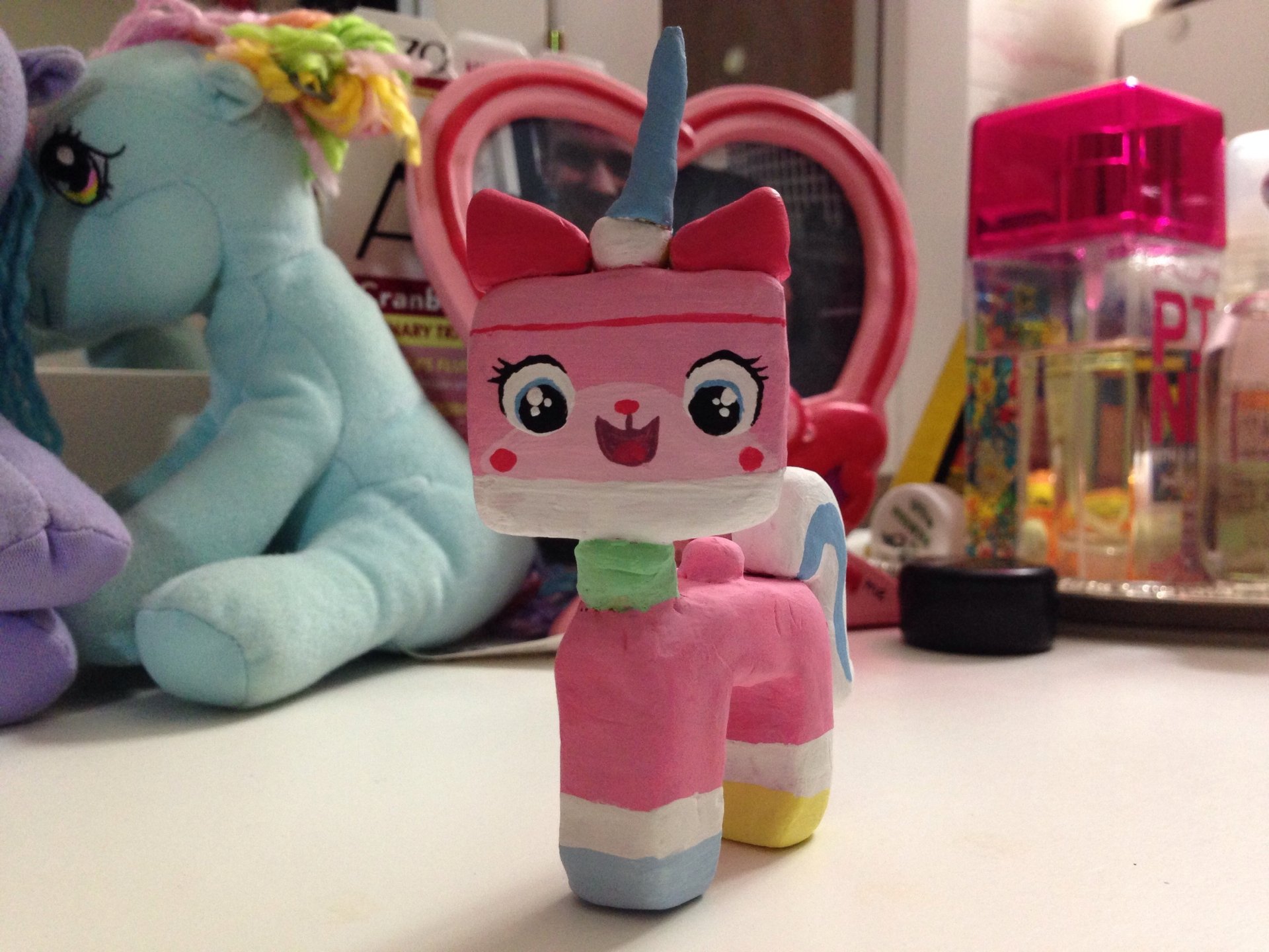 Pink Unikitty toy from The Lego Movie on a desk with plushies and trinkets in soft focus — 2K Quad HD PC desktop wallpaper/background.