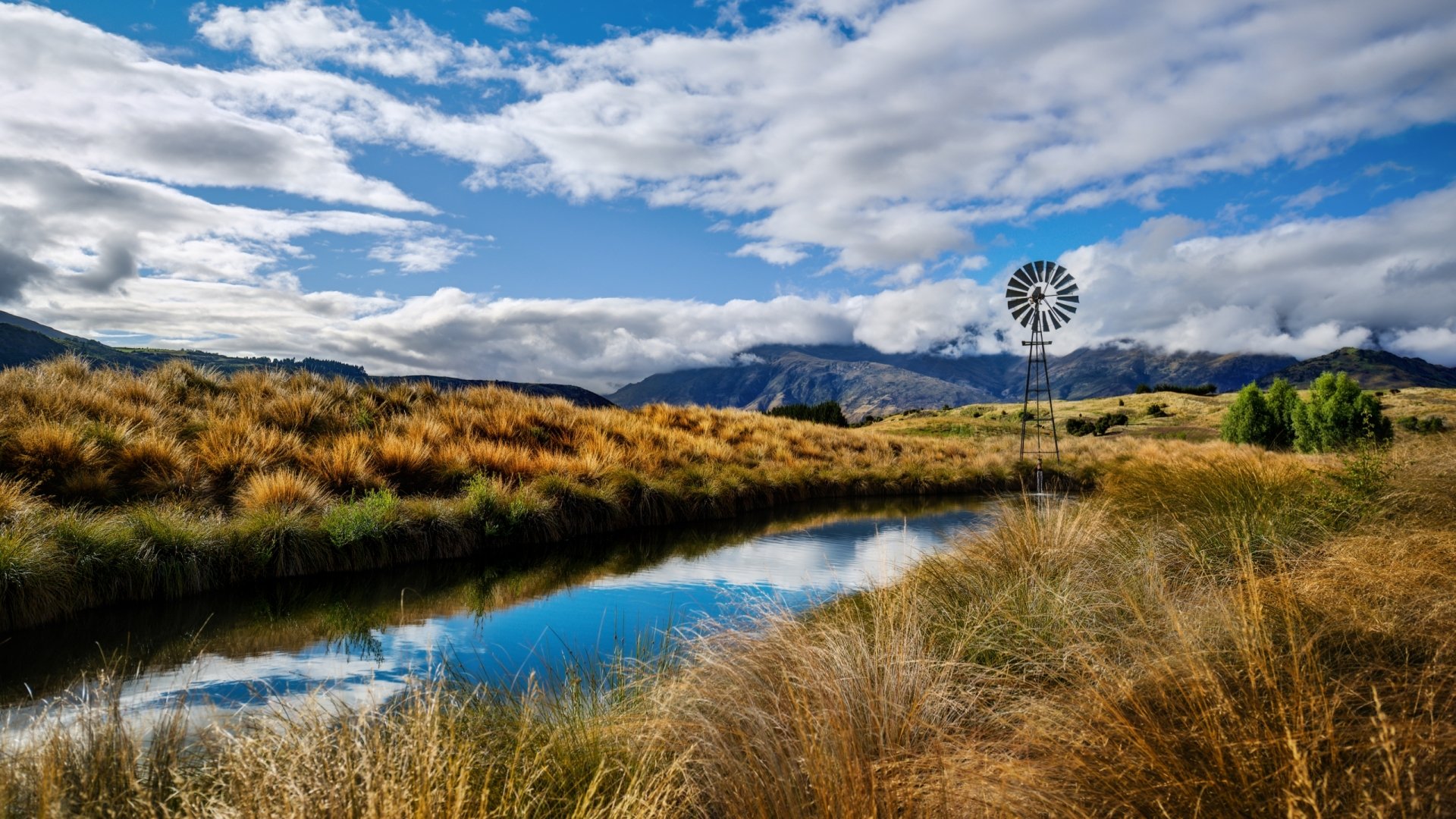 HD PC desktop wallpaper showing a man-made windmill beside a reflective waterway, set in a grassy landscape under a blue sky with scattered clouds.