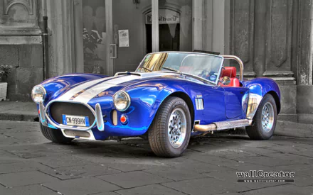 HD PC desktop wallpaper featuring a classic blue AC Cobra vehicle with white racing stripes parked on a cobblestone street in front of old stone buildings.