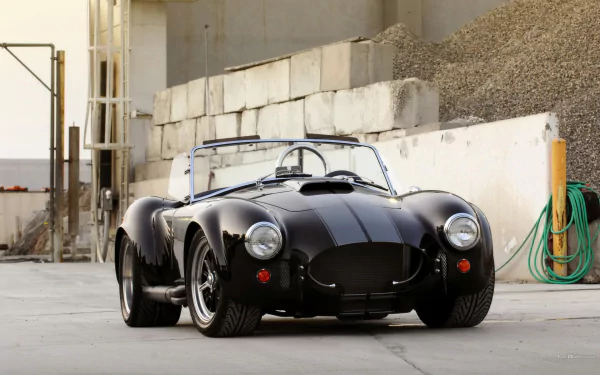 HD PC desktop wallpaper of a sleek black AC Cobra parked in an industrial setting, showcasing its classic design and powerful presence.