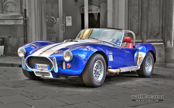 HD PC desktop wallpaper featuring a classic blue AC Cobra vehicle with white racing stripes parked on a cobblestone street in front of old stone buildings.
