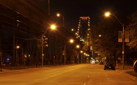 Night view of the Ambassador Bridge from a quiet city street, lit by rows of streetlights and the bridge’s illuminated towers — HD desktop wallpaper.
