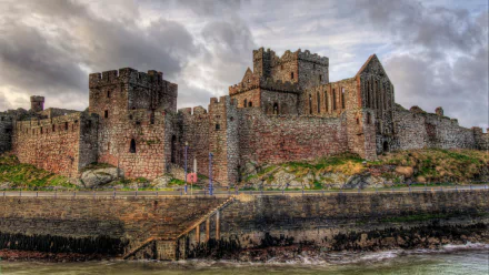 2K Quad HD desktop wallpaper of Peel Castle, a man-made stone fortress on a rocky shore, its towers and battlements beneath dramatic clouds, reflected above choppy harbor waters.