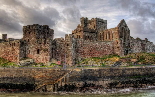 2K Quad HD desktop wallpaper of Peel Castle, a man-made stone fortress on a rocky shore, its towers and battlements beneath dramatic clouds, reflected above choppy harbor waters.