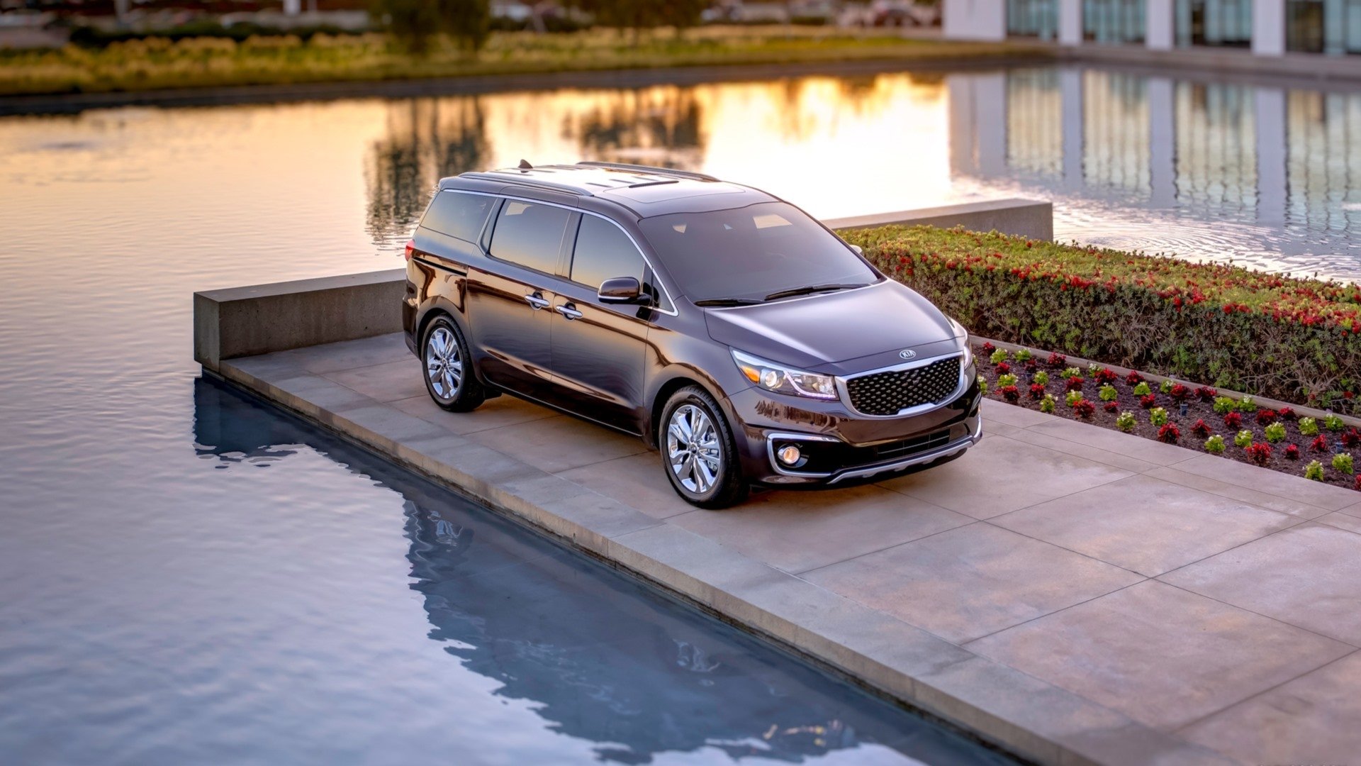 HD PC desktop wallpaper: 2015 Kia Sedona minivan parked on a concrete pier beside reflective water with modern buildings and landscaped beds in the background.