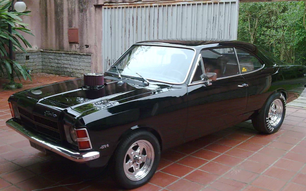 HD desktop wallpaper featuring a classic black Chevrolet Opala Comodoro parked on a tiled patio beside a brick wall and green plants.