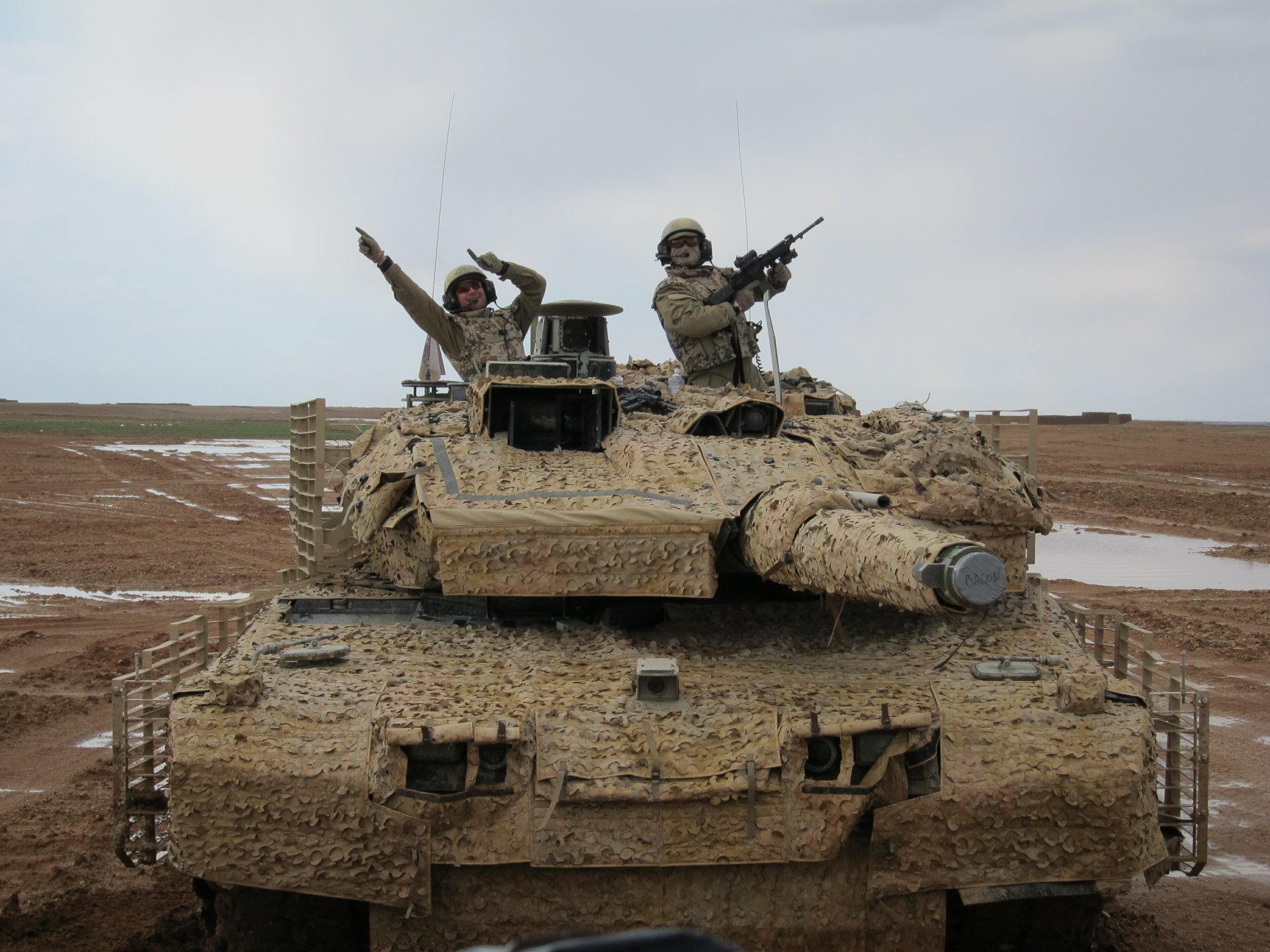 2K Quad HD PC desktop wallpaper and background: mud-splattered military tank front view with two armed soldiers on its turret in a muddy field beneath an overcast sky.