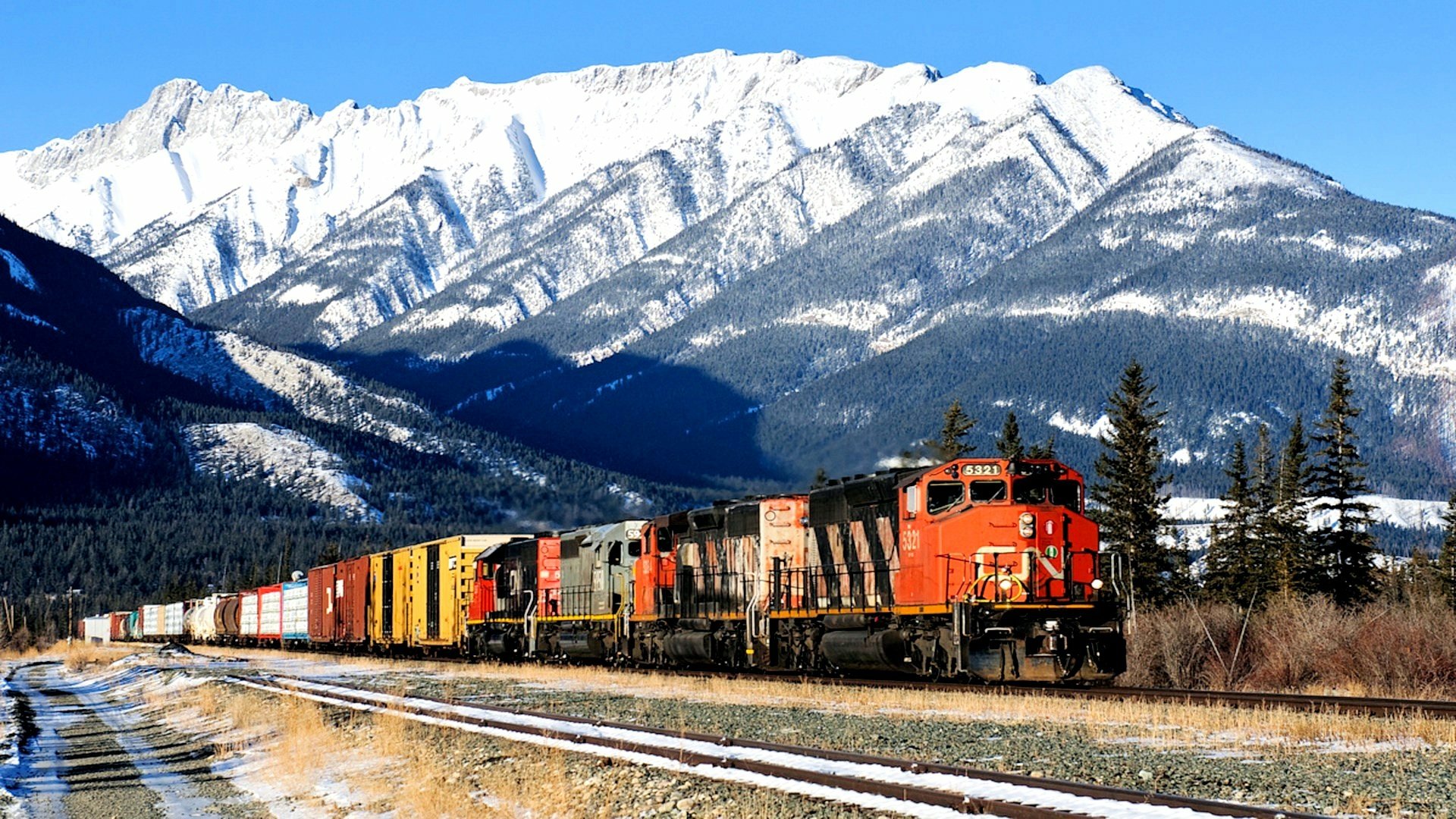 HD desktop wallpaper showing a long freight train traveling through a forested area with snow-covered mountains in the background under a clear blue sky.