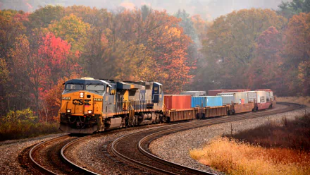 HD desktop wallpaper of a freight train curving along tracks surrounded by autumn trees with vibrant fall foliage under a misty sky.