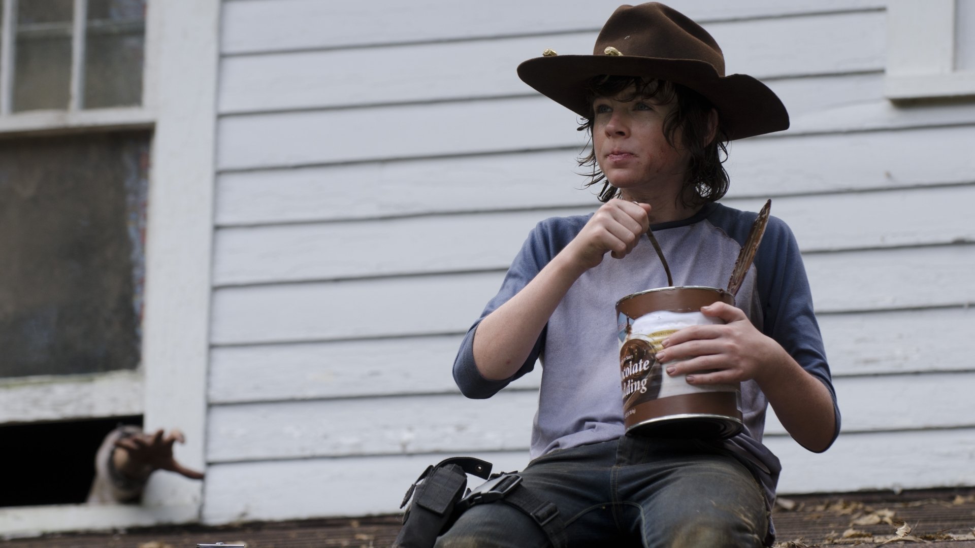 A scene from The Walking Dead featuring a young character in a cowboy hat, sitting on a porch and eating from a container, surrounded by a desolate setting. HD desktop wallpaper.
