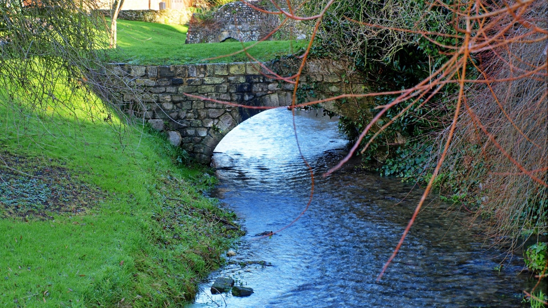 HD desktop wallpaper showcasing a serene stream flowing beneath a stone bridge, surrounded by lush green grass and trees in a natural setting.