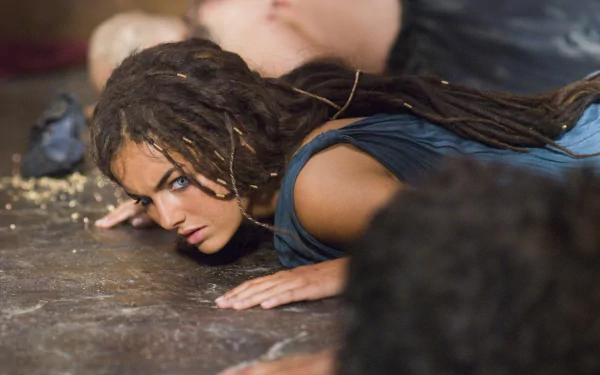 A dramatic scene from the movie 10,000 BC, featuring a young woman with braided hair, intensely focused as she lies on the ground, set against a blurred background. HD wallpaper.
