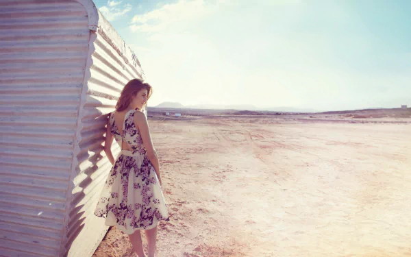 HD PC desktop wallpaper featuring a woman in a floral dress standing beside a corrugated metal wall in a vast, sunlit desert landscape.
