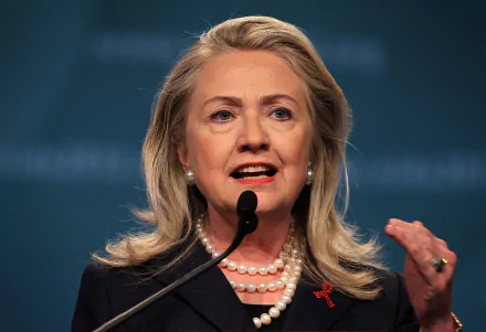 Hillary Rodham Clinton speaking passionately at a podium, adorned with pearls and a red ribbon, set against a soft blue background. A striking HD wallpaper featuring a prominent celebrity.