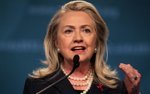 Hillary Rodham Clinton speaking passionately at a podium, adorned with pearls and a red ribbon, set against a soft blue background. A striking HD wallpaper featuring a prominent celebrity.