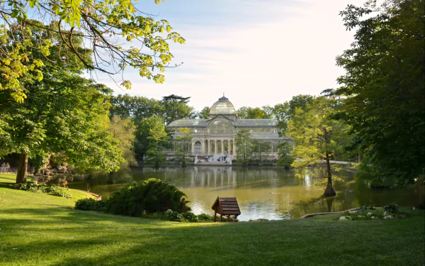 HD desktop wallpaper showing the Palacio de Cristal, a man-made glass and metal structure surrounded by lush greenery and a reflective pond in a serene park setting.