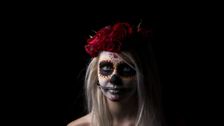 Artistic sugar skull portrait of a pale woman with painted skull makeup and a red rose crown on a black background — 8K Ultra HD PC desktop wallpaper and background.