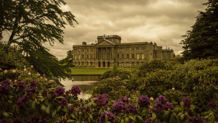 Man-made Lyme Park mansion by a lake, framed by purple rhododendrons beneath a moody sky — 2K Quad HD PC desktop wallpaper and background.