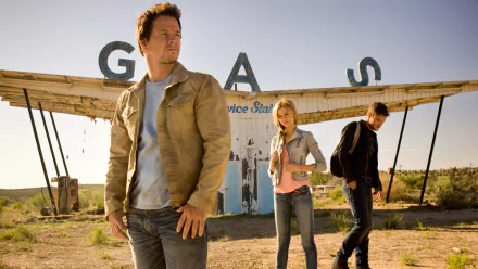 A scene from Transformers: Age of Extinction featuring three characters in front of an abandoned gas station, conveying a sense of adventure in a post-apocalyptic landscape.