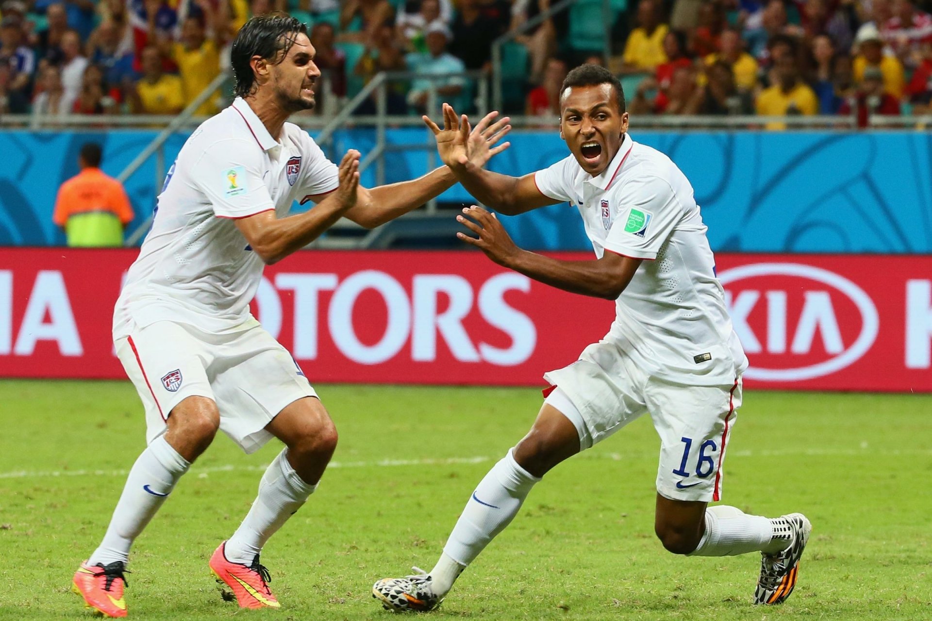 Two soccer players in white jerseys celebrate on the field, with a stadium and advertising boards in the background. This HD wallpaper captures the excitement of a live soccer match.