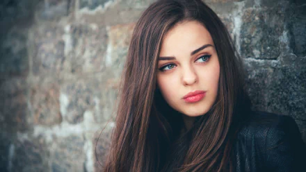 HD desktop wallpaper of a woman with long brown hair, gazing thoughtfully to the side, standing against a textured stone wall.