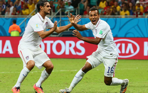 Two soccer players in white jerseys celebrate on the field, with a stadium and advertising boards in the background. This HD wallpaper captures the excitement of a live soccer match.