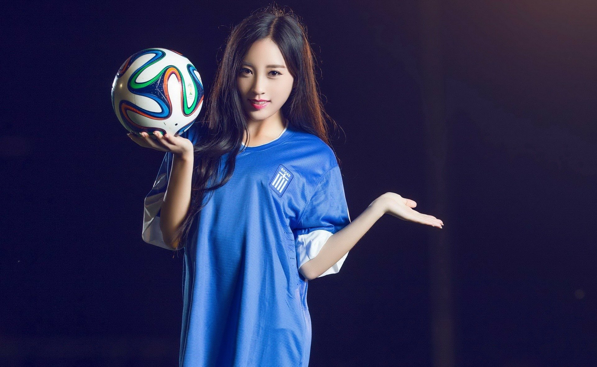 Asian woman in a Greece soccer jersey holding a colorful football, set against a dark background in an HD desktop wallpaper.