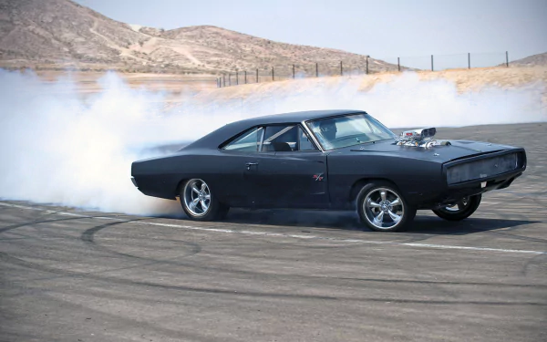 A sleek black Dodge Charger R/T drifts on a dusty track, with tires smoking against a backdrop of rolling hills, showcasing its powerful stance and dynamic performance. HD desktop wallpaper.