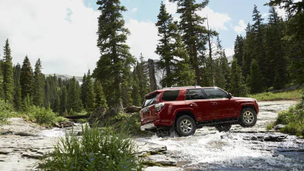 A red Toyota 4Runner 4x4 SUV drives through a shallow stream surrounded by tall pine trees in a forest, captured in this HD desktop wallpaper.