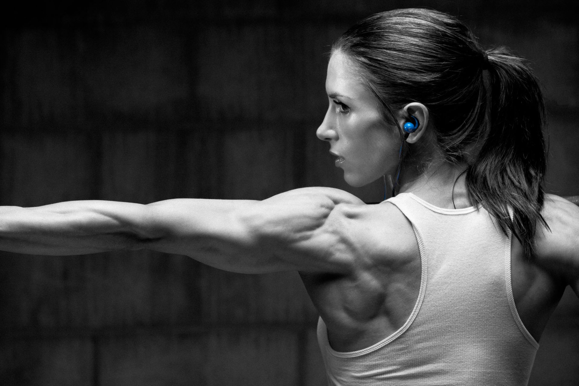 HD desktop wallpaper of a fit woman in a sports tank top stretching her arm, showcasing toned muscles and wearing blue earphones against a dark background.