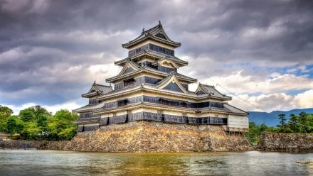 HD desktop wallpaper showcasing Matsumoto Castle, a historic man-made Japanese fortress surrounded by water under a dramatic cloudy sky.