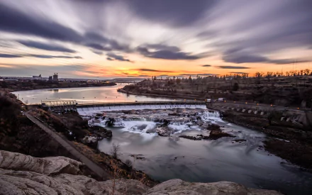 A stunning HD desktop wallpaper showcasing a dam at sunset, with flowing water and dynamic cloud patterns, framed by rocky terrain and a serene river landscape.