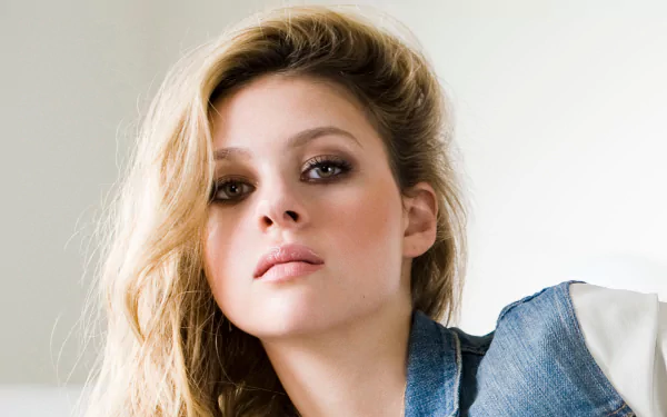A striking HD wallpaper featuring American actress Nicola Peltz, showcasing her stylish look with tousled hair and a denim jacket against a softly lit background.