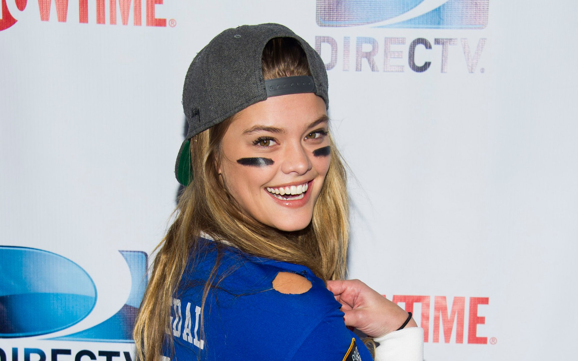 Danish model and celebrity Nina Agdal smiles at a media event, wearing a backward cap and sports face paint, captured in an HD desktop wallpaper.