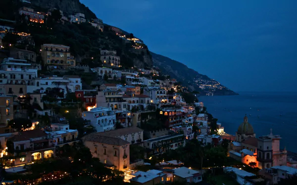 Night view of the man-made coastal town of Positano, Salerno, Italy, with illuminated buildings cascading down the hillside against a deep blue sky and sea.
