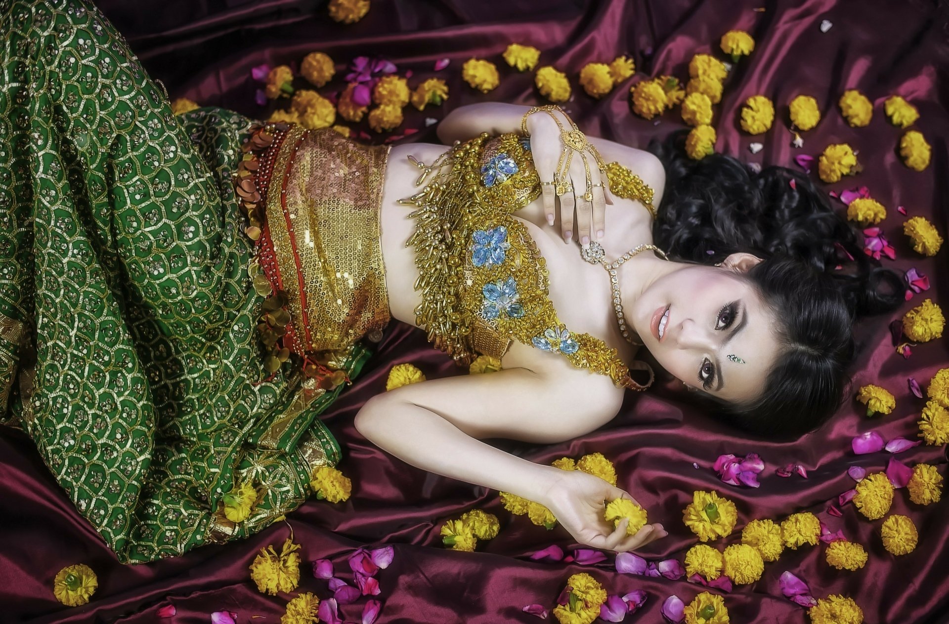 HD desktop wallpaper featuring an Asian woman in traditional Thai attire, lying on a purple silk fabric adorned with yellow marigold flowers, showcasing oriental beauty.
