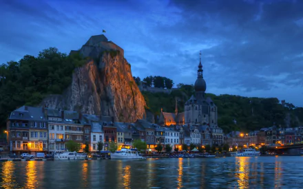 A stunning night view of Notre Dame de Dinant, Belgium, showcasing illuminated houses along the river with a dramatic rock formation in the background, creating a captivating urban landscape.