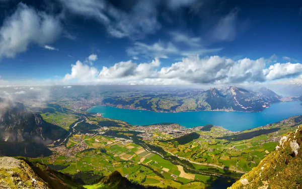 A breathtaking Swiss landscape with vibrant green valleys, a large lake, and dramatic clouds under a bright blue sky, captured in HD photography for a desktop wallpaper.