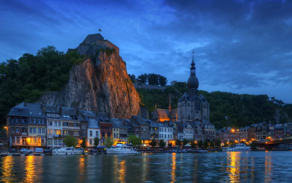 A stunning night view of Notre Dame de Dinant, Belgium, showcasing illuminated houses along the river with a dramatic rock formation in the background, creating a captivating urban landscape.