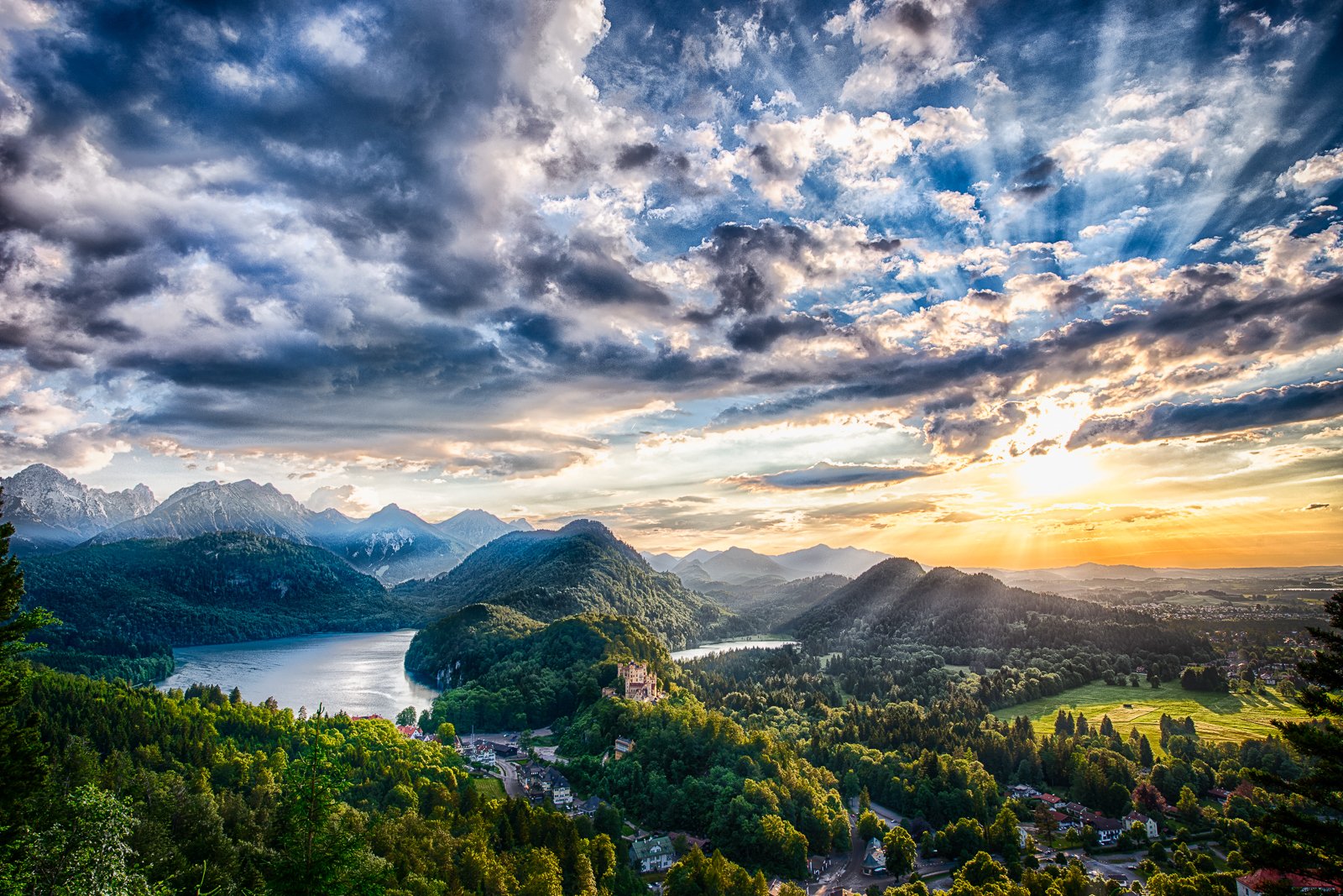 A stunning HD desktop wallpaper featuring Neuschwanstein Castle surrounded by lush greenery and mountains under a dramatic sky at sunset.