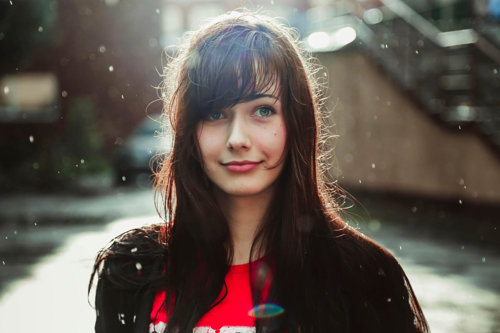 A woman with long dark hair smiles softly in a natural outdoor setting, illuminated by soft light and surrounded by gentle raindrops. This image serves as an HD desktop wallpaper and background.