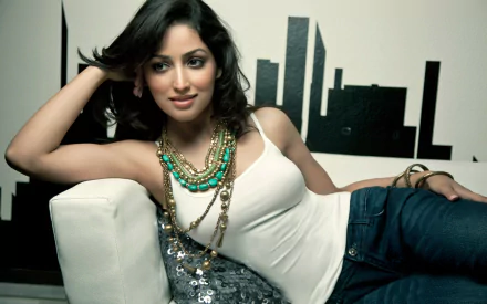 A stylish woman, Yami Gautam, poses elegantly on a couch, wearing a white top and layered necklaces, with a city skyline backdrop, captured in an HD desktop wallpaper.