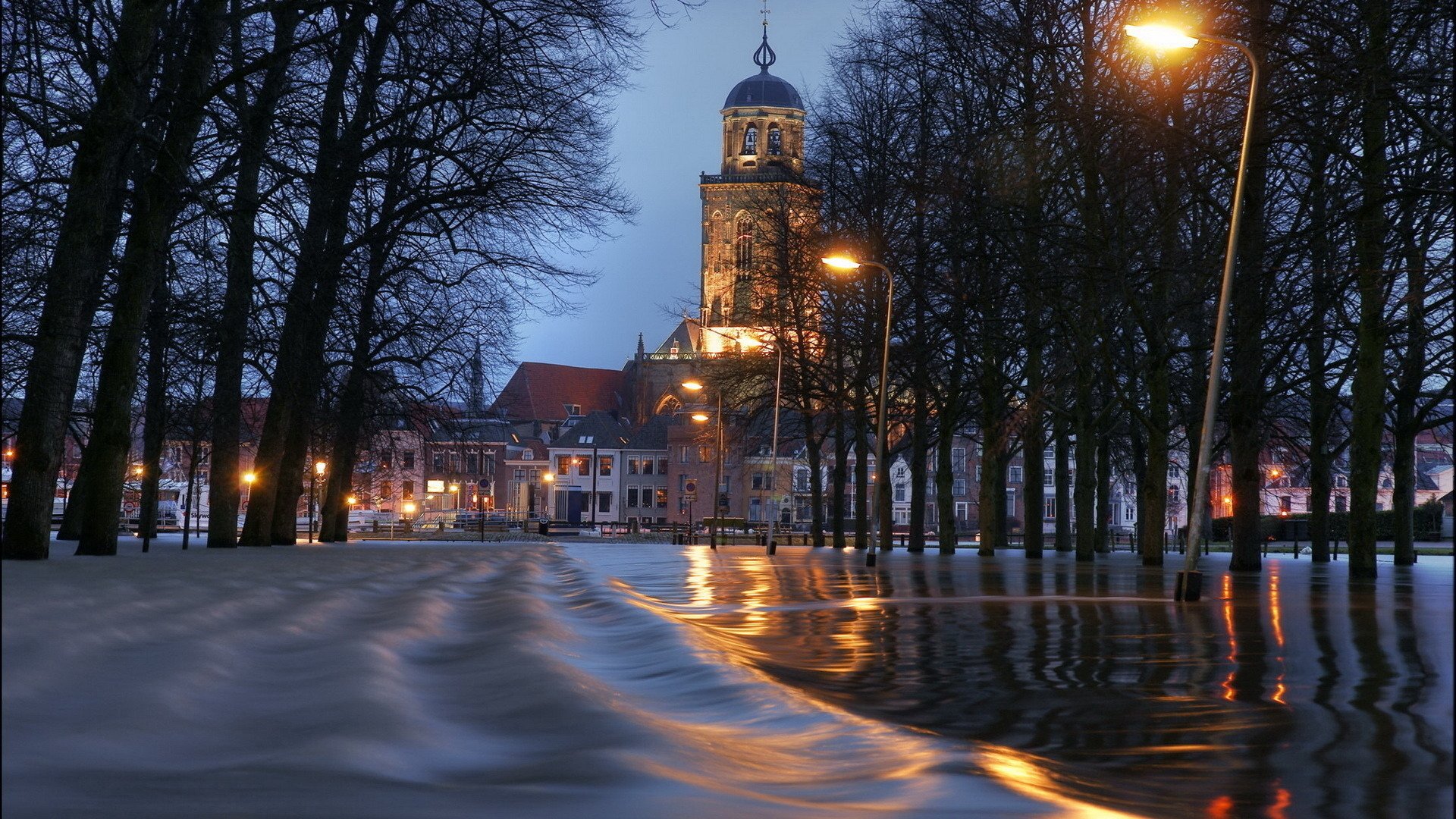 HD desktop wallpaper: evening view of the religious Lebuinus Church in the Netherlands framed by leafless trees, wet reflective cobblestone path and warm streetlights.