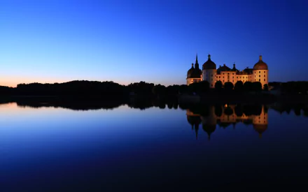 Man-made Moritzburg Castle at dusk, lit and silhouetted against a deep blue sky, reflected in a calm lake — 2K Quad HD PC desktop wallpaper/background.