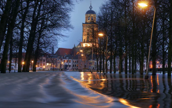 HD desktop wallpaper: evening view of the religious Lebuinus Church in the Netherlands framed by leafless trees, wet reflective cobblestone path and warm streetlights.