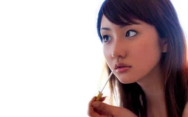 An Asian woman with long hair engages in a sensual moment, holding a lip gloss wand. The soft background creates a serene atmosphere, making this an elegant HD desktop wallpaper.