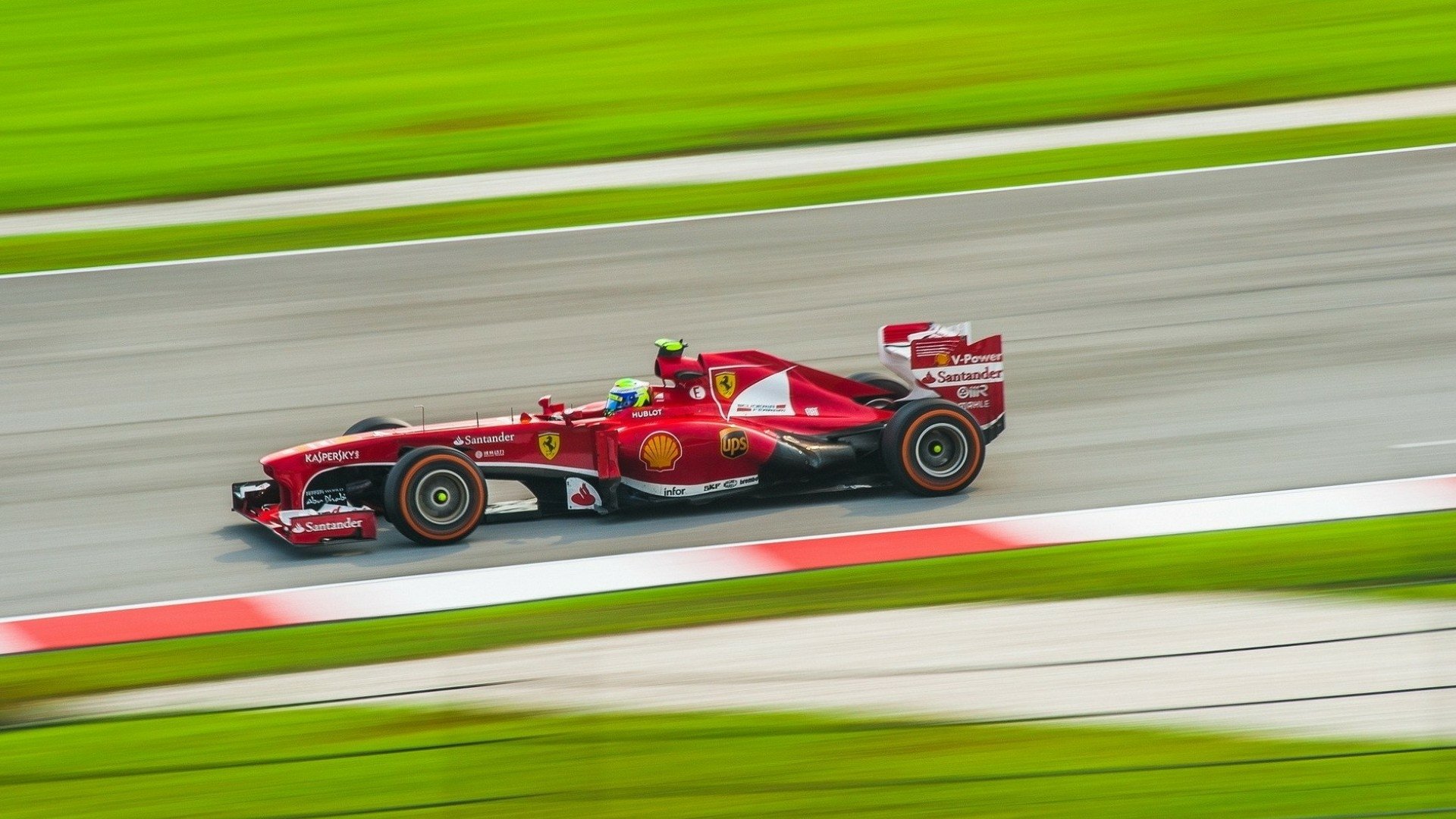 HD PC desktop wallpaper showing a red Ferrari F1 race vehicle speeding along a racetrack with motion-blurred green runoff and asphalt streaks.