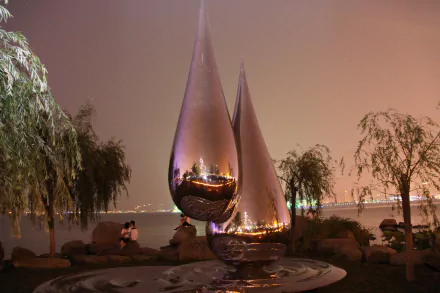 Nighttime view of reflective, teardrop-shaped sculptures by the waterfront in Suzhou, China, surrounded by trees and city lights in the background.