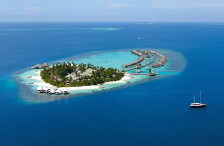 Aerial view of a tropical Maldives atoll with lush village island, overwater hotel bungalows, and a ship sailing in the deep blue sea under clear skies.